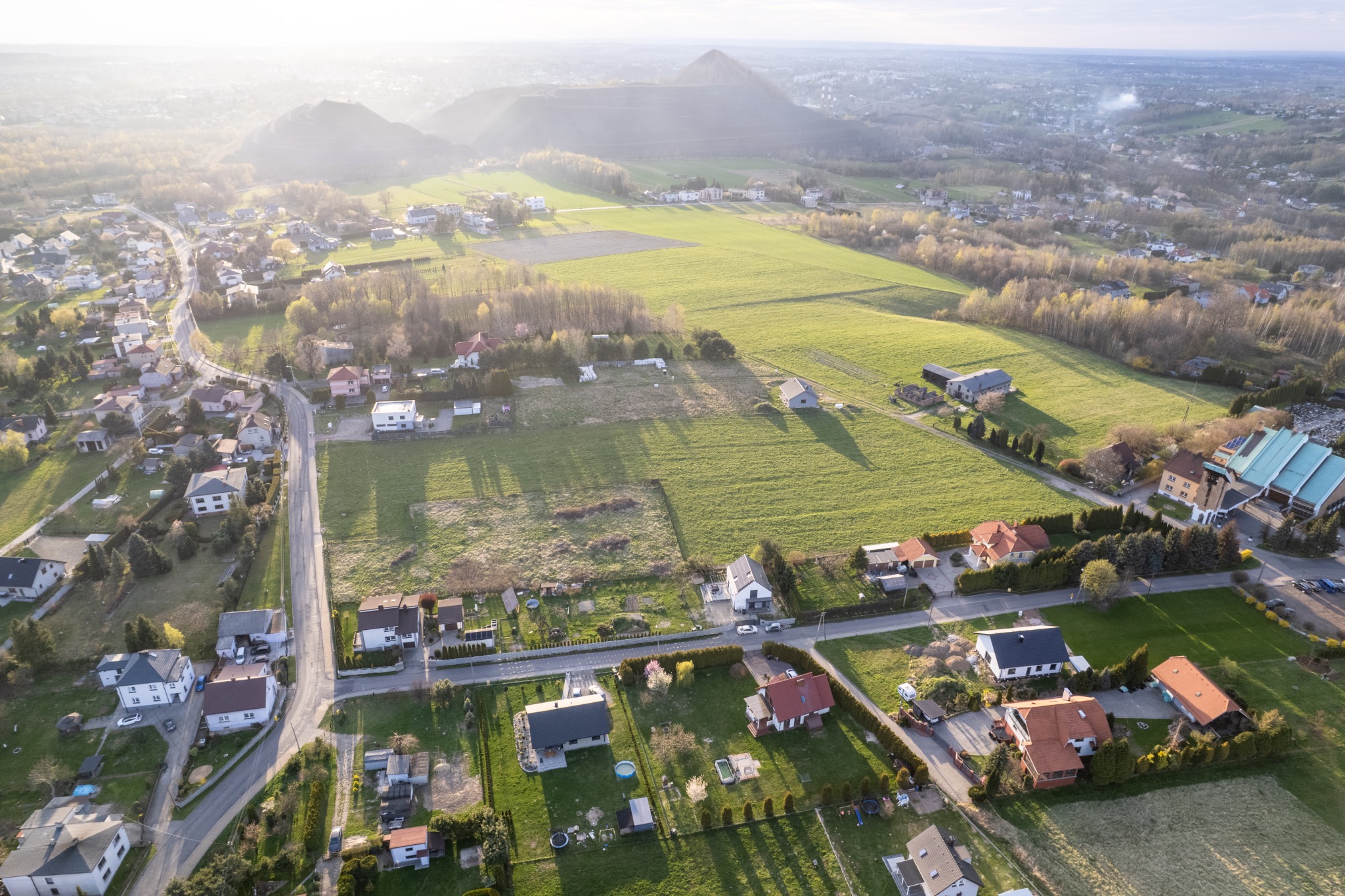 Panoramiczny widok z lotu ptaka na wiejską okolicę z domami, polami i w oddali wzniesieniem, oświetlony ciepłym światłem poranka.