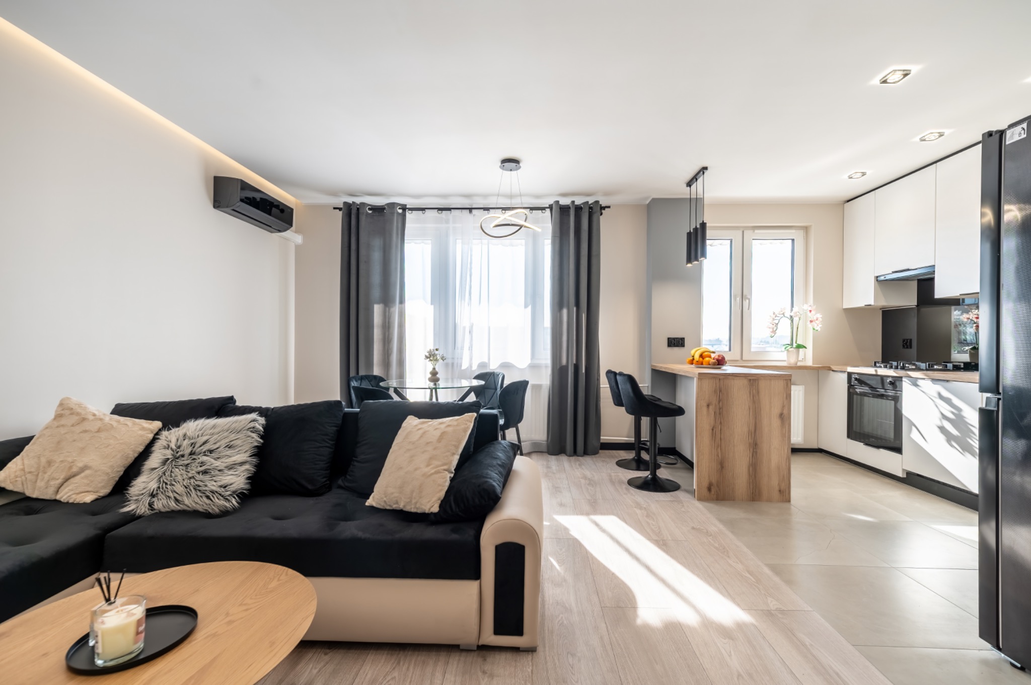 Jasne wnętrze mieszkania z aneksem kuchennym, czarna sofa z dekoracyjnymi poduszkami, okrągły stół z krzesłami, wyspa kuchenna z hokerami i nowoczesne oświetlenie.
