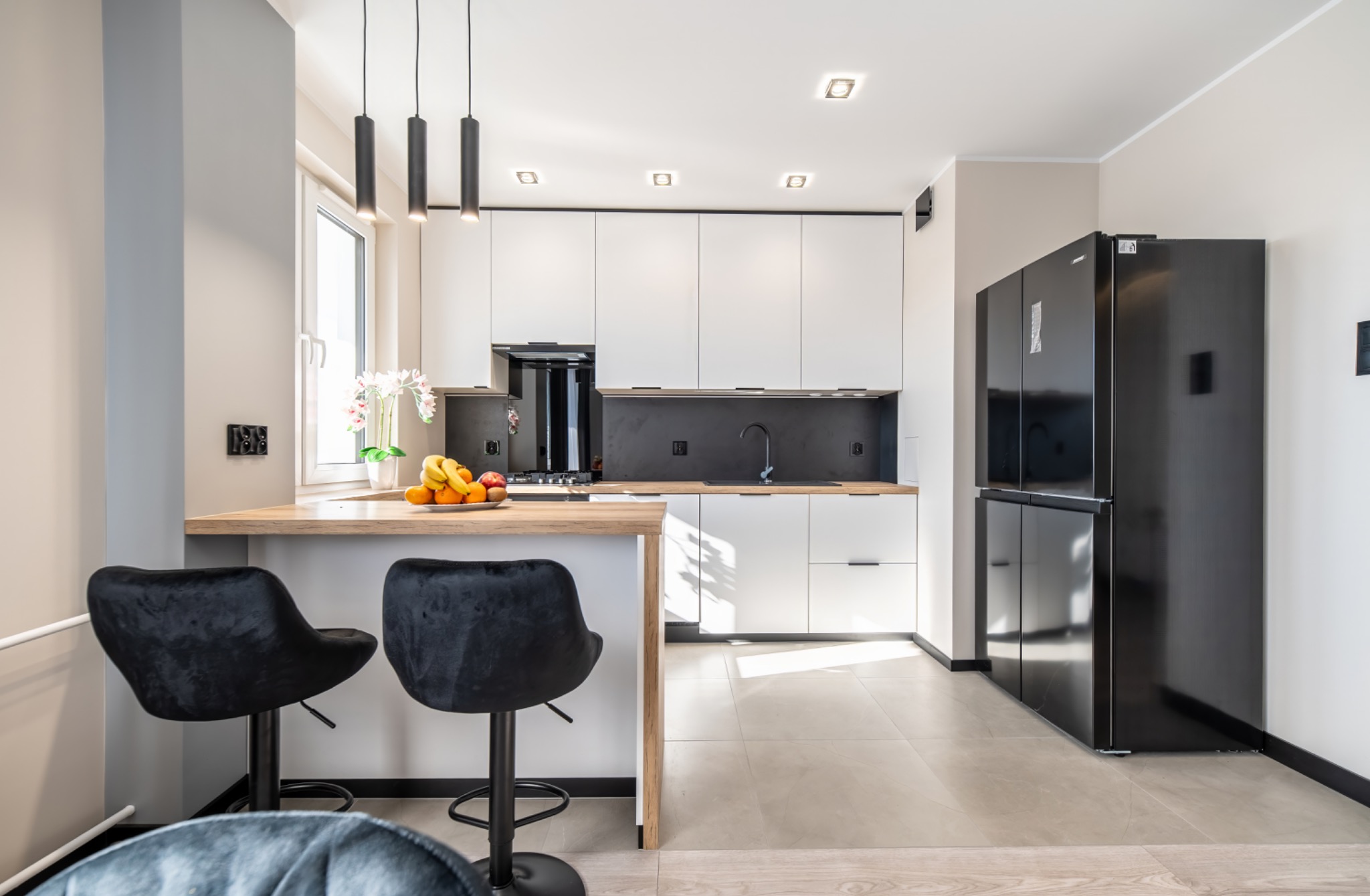 Nowoczesna kuchnia z białymi szafkami, czarnym panelem nad blatem, drewnianym blatem wyspowym i czarną lodówką side-by-side; na blacie owoce w misce, nad blatem trzy czarne lampy wiszące.