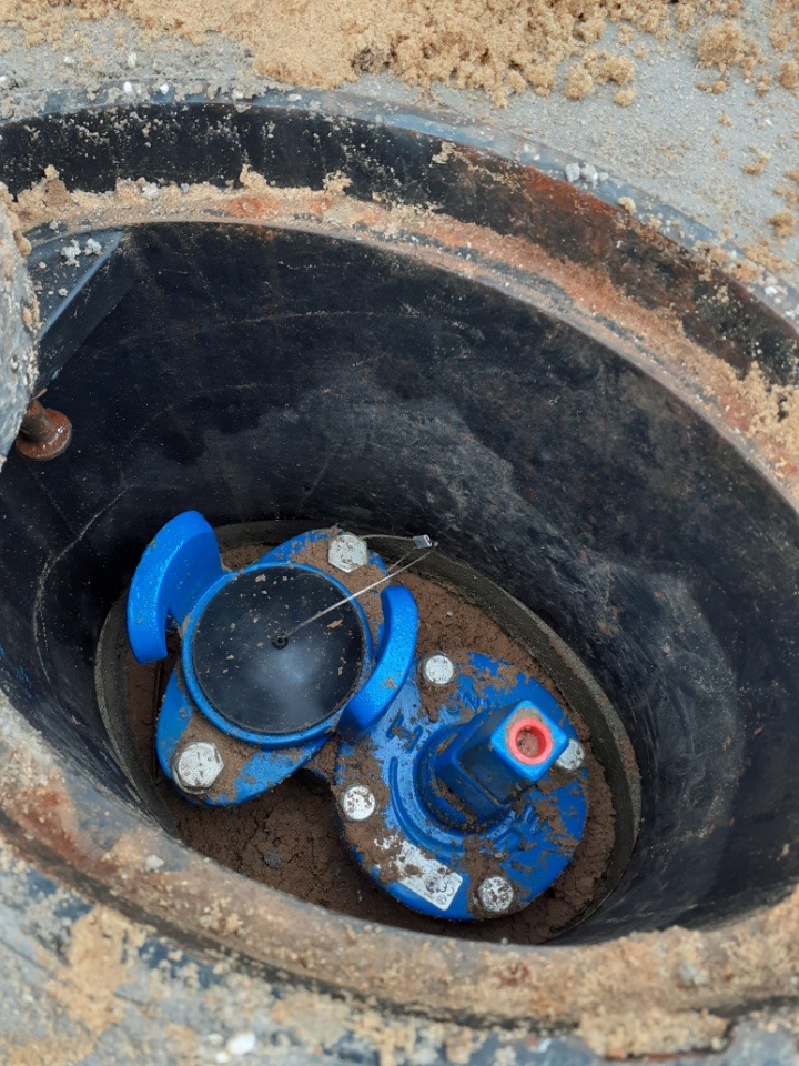 Niebieski zawór wodny w czarnej studzience, widoczne ślady ziemi i piasku na zaworze i w studzience.