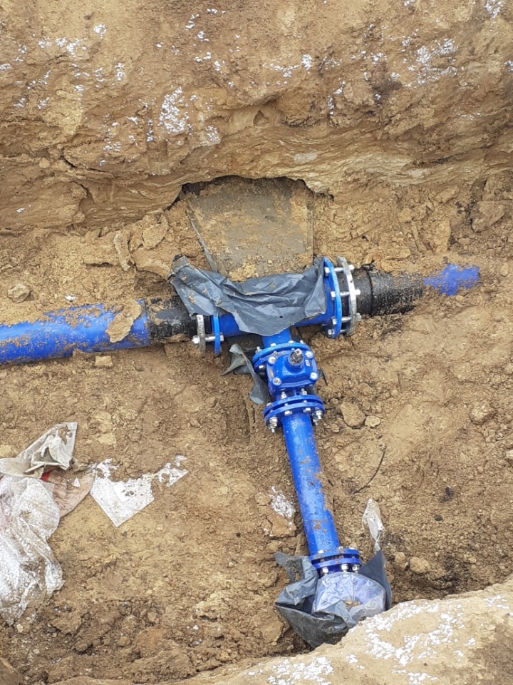 Niebieskie rury połączone kształtką w wykopie ziemnym, zabezpieczone folią, widoczne ślady prac ziemnych.