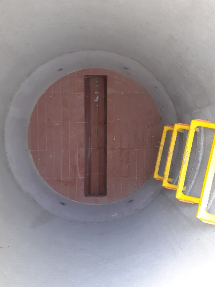 Wnętrze betonowej studni kanalizacyjnej z ceglanym dnem i drabinką ze szczeblami w żółtej osłonie.