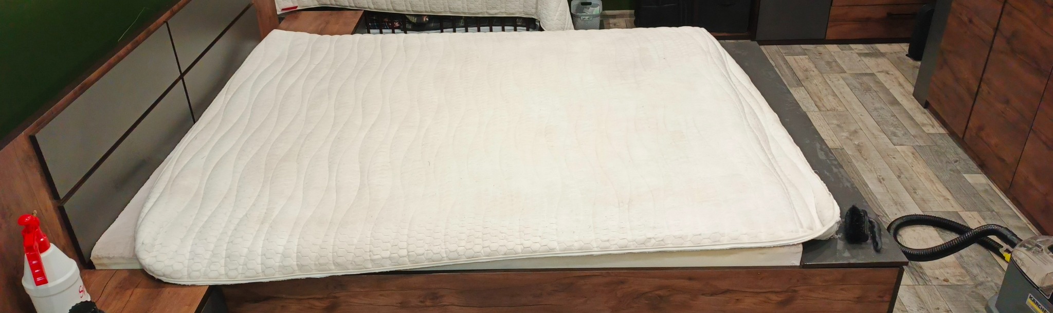 Materac z widocznymi plamami, częściowo leżący na łóżku, obok spryskiwacz i odkurzacz piorący Karcher z czarnym wężem na podłodze w panelach.