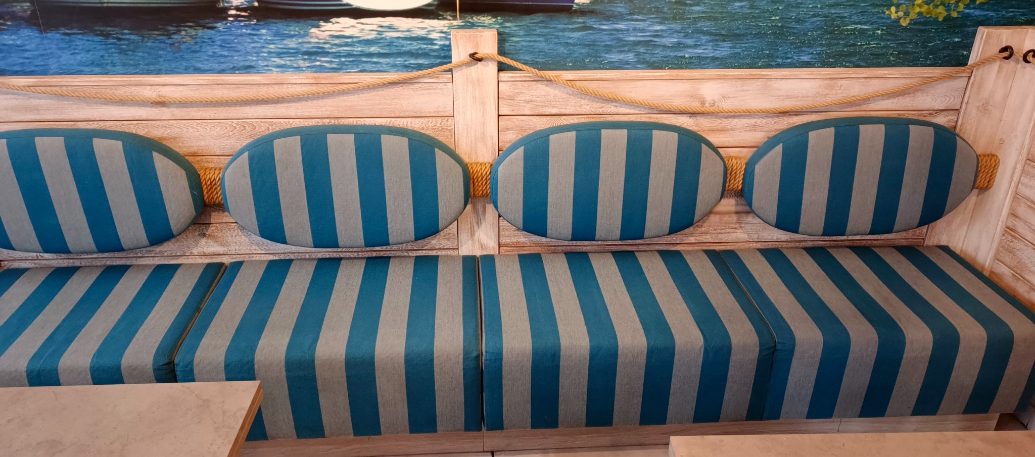Tapicerowana kanapa w stylu marynistycznym z niebiesko-szarymi pasami, oparcie z czterech owalnych poduszek, całość na tle fototapety z motywem morza i łodzi.