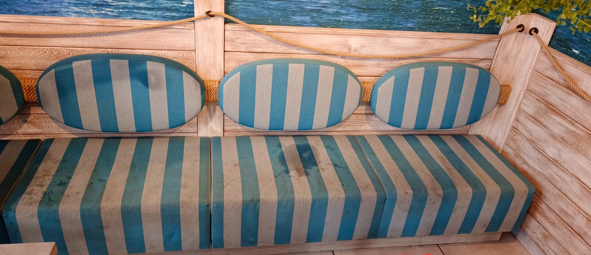 Tapicerowana kanapa w stylu marynistycznym z niebiesko-białymi pasami, widoczne plamy na siedzisku, drewniana konstrukcja z linami i tłem imitującym morze.