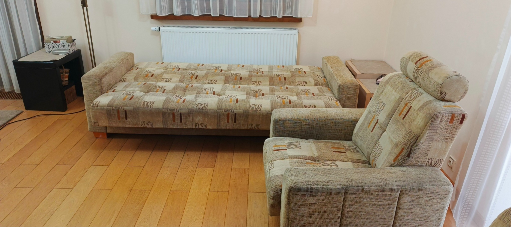 Rozłożona sofa i fotel tapicerowane beżową tkaniną w geometryczne wzory, drewniana podłoga, biały grzejnik na ścianie w tle.