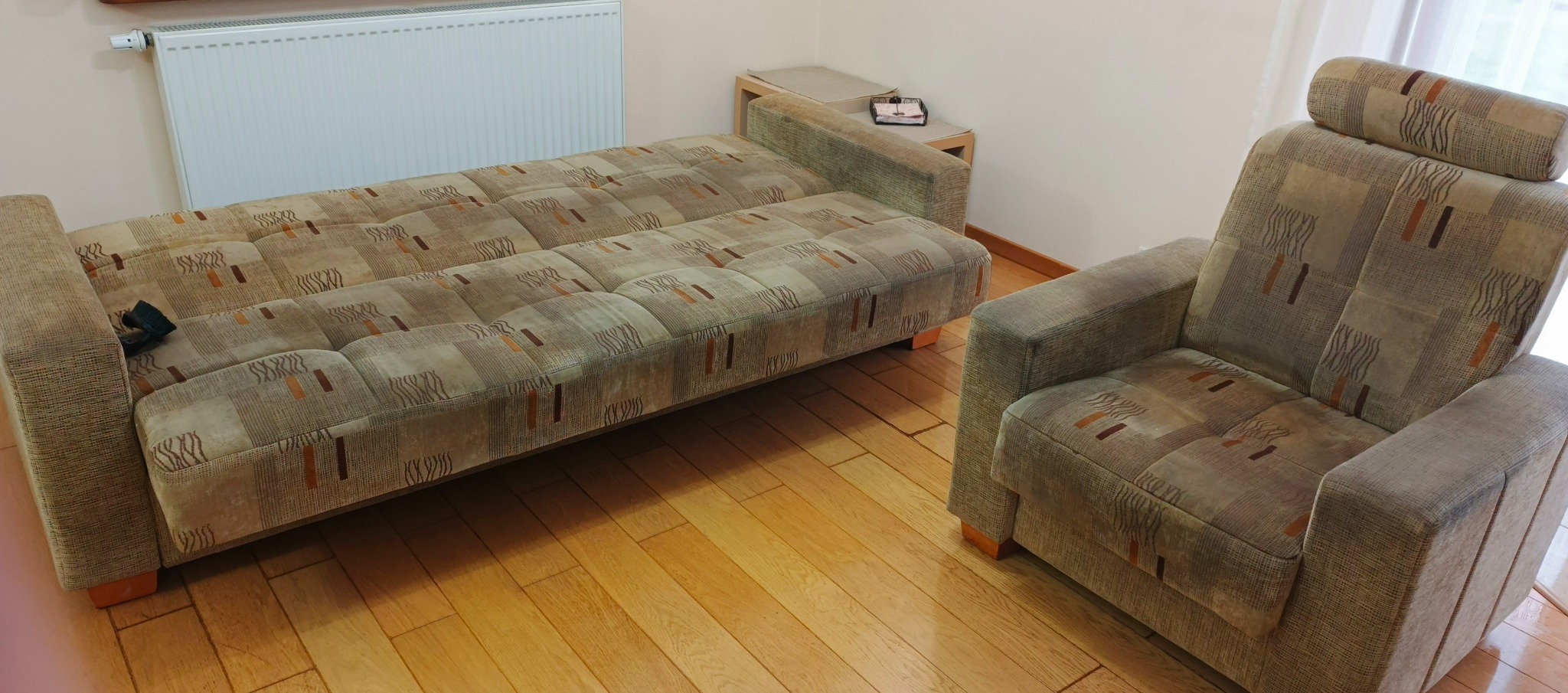 Rozłożona kanapa i fotel tapicerowane w geometryczny wzór w odcieniach beżu i brązu, stojące na drewnianej podłodze w pokoju z grzejnikiem i stolikiem pomocniczym.