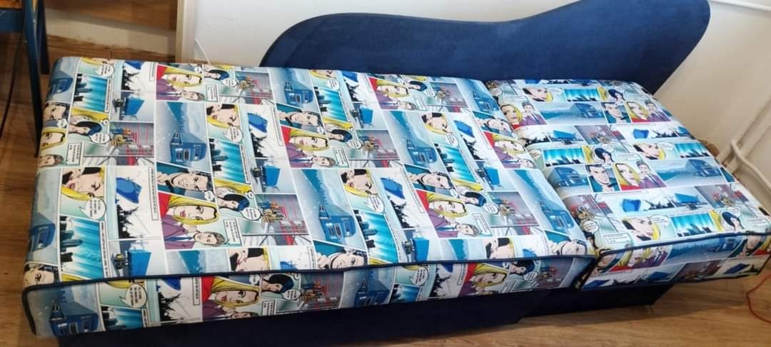 Rozłożona sofa z ciemnoniebieskim obiciem i wzorem w komiksowe kadry z postaciami i budynkami, widok z góry.