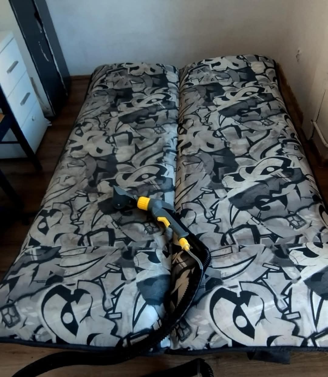Dysza parowa czyszcząca materac z szarym wzorem graffiti, widoczny fragment węża i obudowy urządzenia.