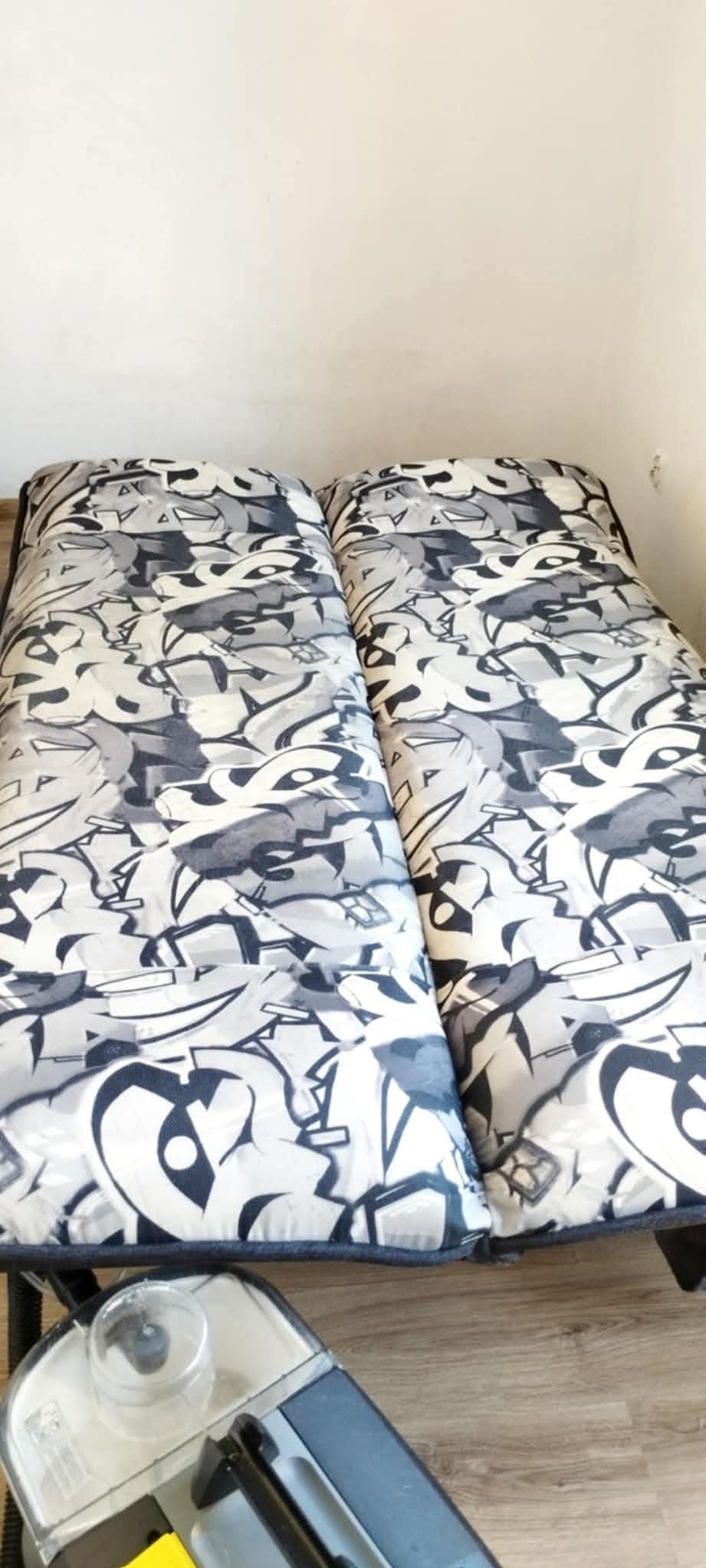 Dwa materace z szaro-białym wzorem graffiti leżące obok siebie, częściowo widoczny odkurzacz piorący na drewnianej podłodze, ściana w tle.