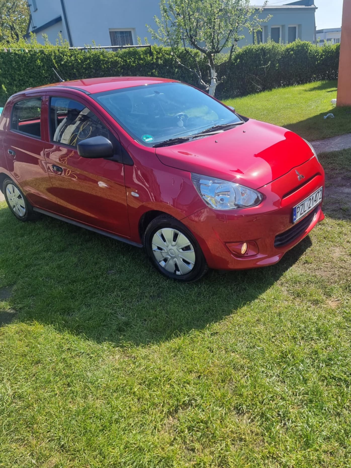 Czerwony samochód osobowy marki Mitsubishi Mirage zaparkowany na trawniku przed domem w słoneczny dzień. Widoczne tablice rejestracyjne.