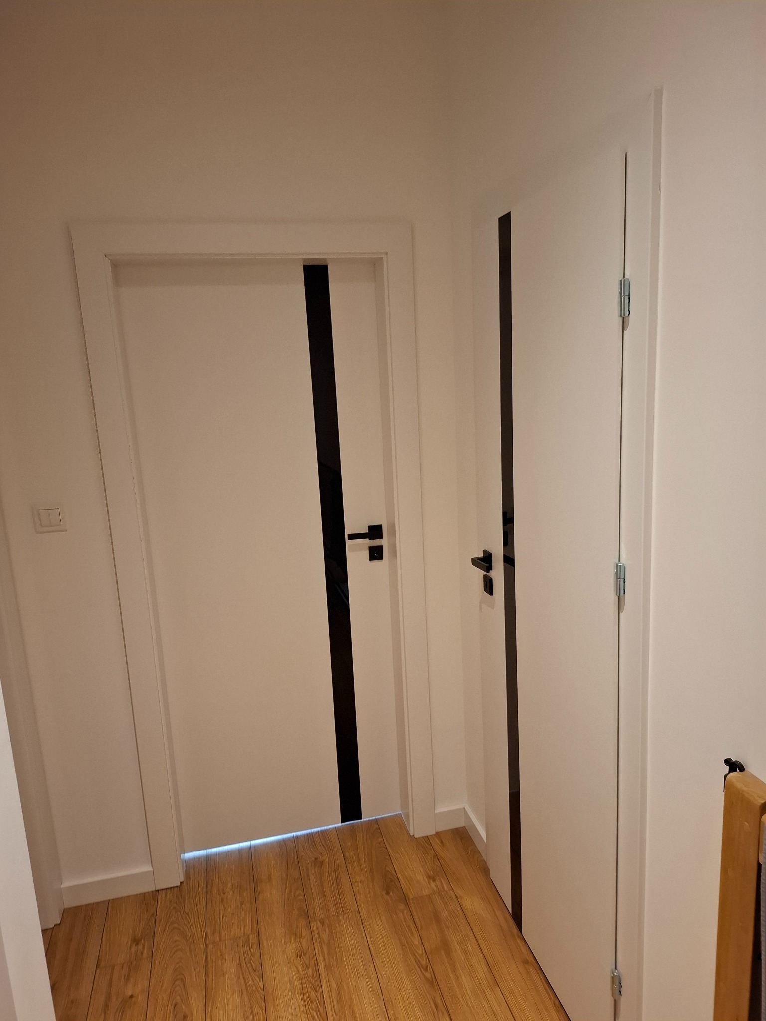 Dwa białe drzwi z czarnymi akcentami w nowoczesnym wnętrzu. Na podłodze panele imitujące drewno. Minimalistyczny design z widocznymi detalami wykończenia.