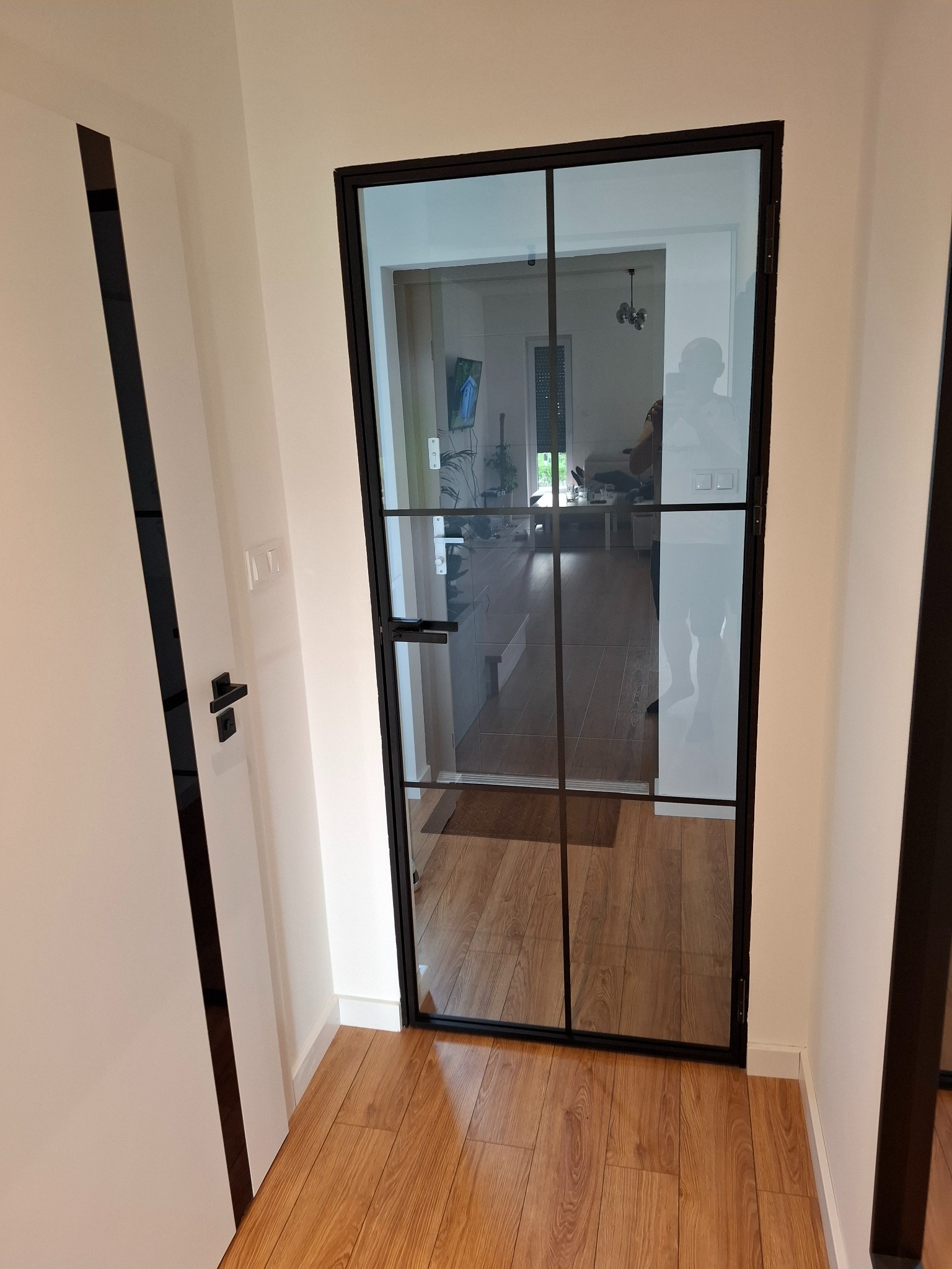 Nowoczesne drzwi szklane z czarną ramą wewnątrz mieszkania. Parkiet na podłodze, widok na salon przez szybę. Minimalistyczny design.