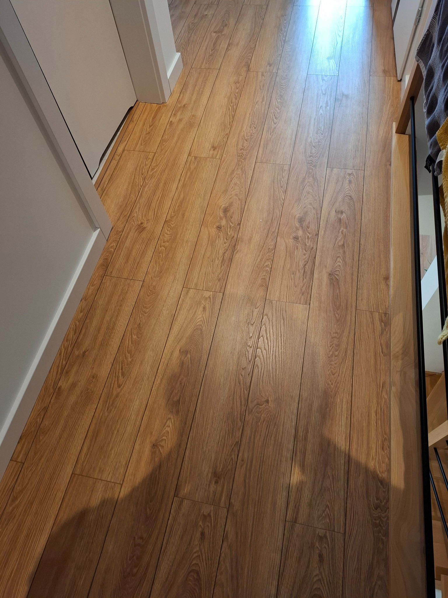 Ułożone panele podłogowe w odcieniach brązu, imitujące drewno, z widoczną strukturą i wzorem słojów, przylegające do białej ościeżnicy drzwiowej.