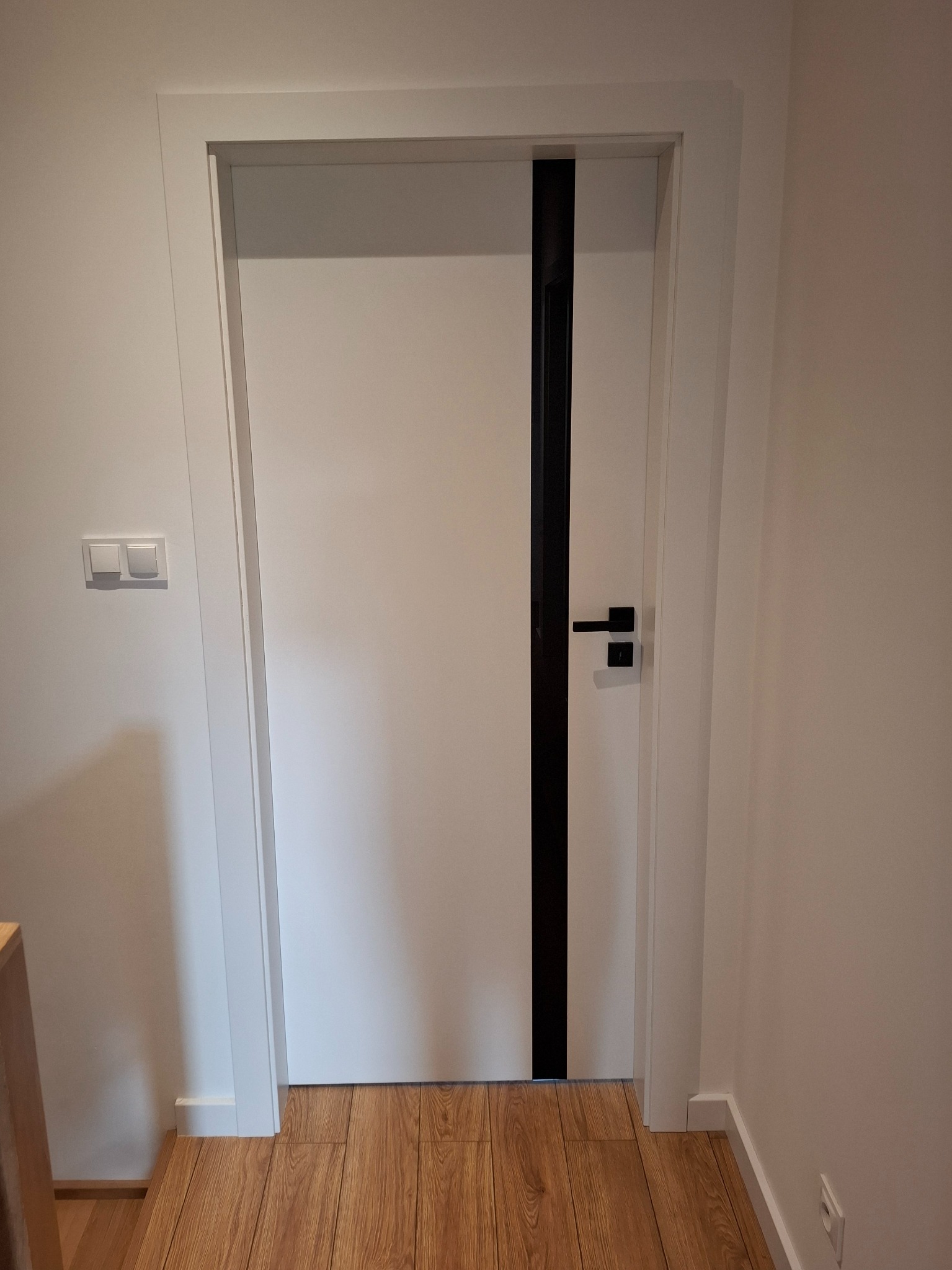 Białe drzwi z czarnym, pionowym akcentem i klamką na tle jasnych ścian i paneli podłogowych. Nowoczesny design, minimalistyczne wykończenie.