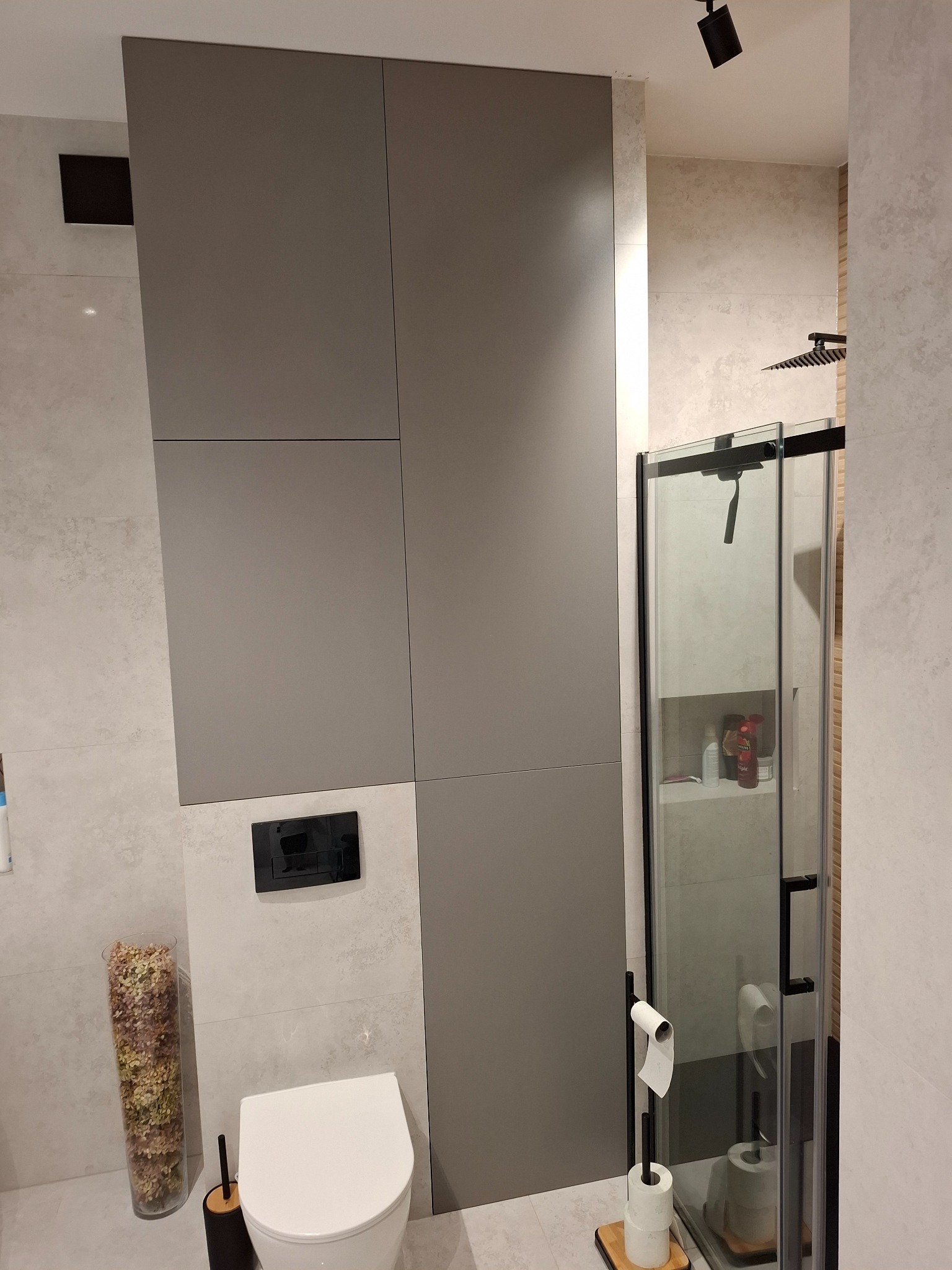 Nowoczesna łazienka z szarą zabudową, podwieszaną toaletą i kabiną prysznicową z czarnymi detalami. Uwagę zwraca minimalistyczny design i stonowana kolorystyka.