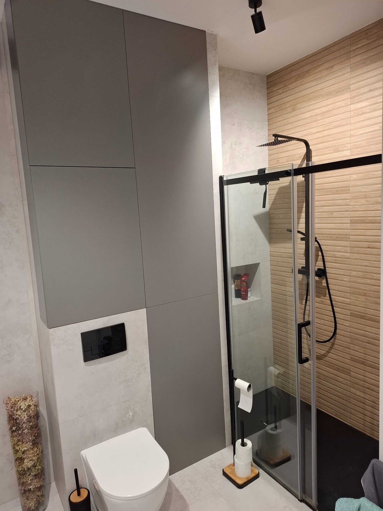 Nowoczesna łazienka z kabiną prysznicową typu walk-in, szarą zabudową meblową i toaletą. Ściany wyłożone jasnymi płytkami, akcent drewnopodobnych płytek pod prysznicem.