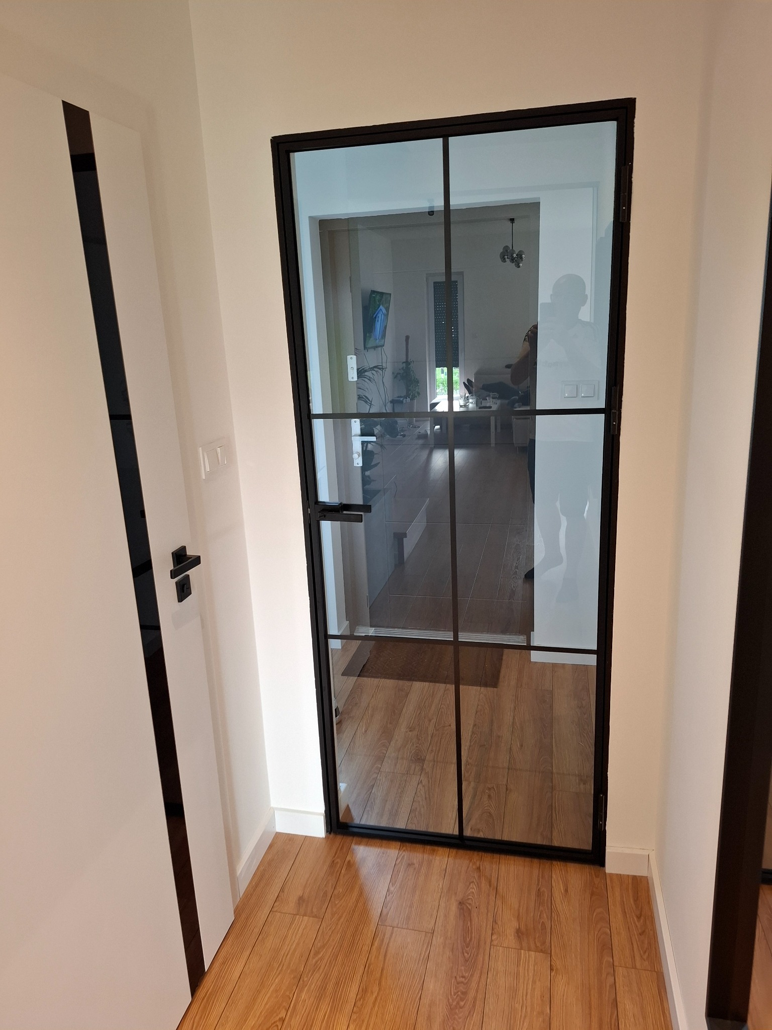 Loftowe drzwi szklane z czarną ramą, widoczne z perspektywy wnętrza, z fragmentem białych drzwi po lewej stronie i drewnianą podłogą.