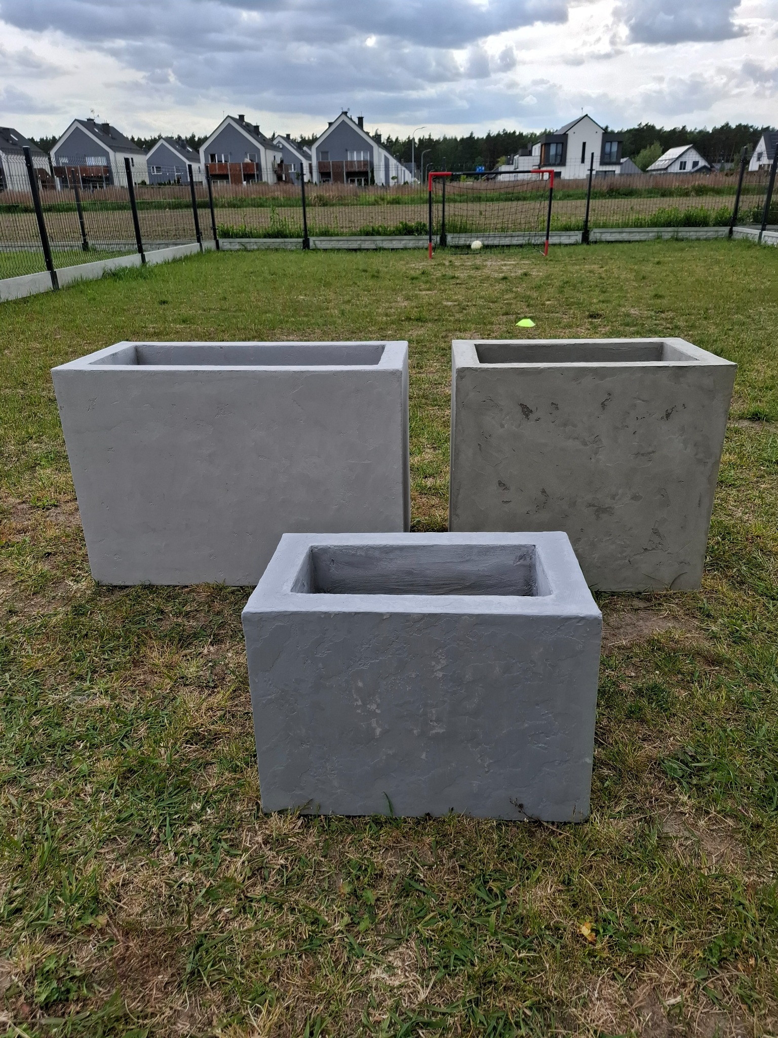 Trzy betonowe donice o różnej wielkości na trawniku przed domem, z widocznym ogrodzeniem i budynkami w tle. Donice mają surową fakturę.