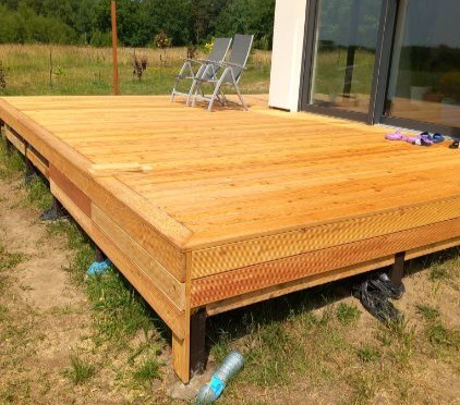 Jasny, drewniany taras z widocznymi śladami montażu, dwie metalowe krzesła w tle, częściowo odsłonięte podpory, trawa i butelka wody na pierwszym planie.