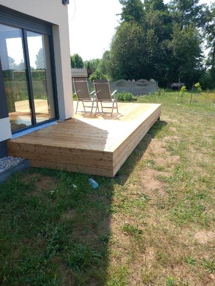 Nowoczesny taras drewniany z dwoma krzesłami, przylegający do budynku z dużym oknem przesuwnym, otoczony zielenią i elementami ogrodzenia.