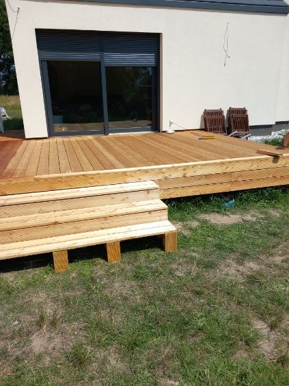 Nowy, drewniany taras z schodami przed domem z dużym oknem. Widoczne krzesła na tarasie i trawa. Jasne drewno, naturalne światło.