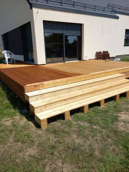 Drewniany taras z jasnymi schodami przed domem z dużym oknem. Część tarasu pomalowana na brązowo, część w naturalnym odcieniu drewna. Trawa wokół tarasu.