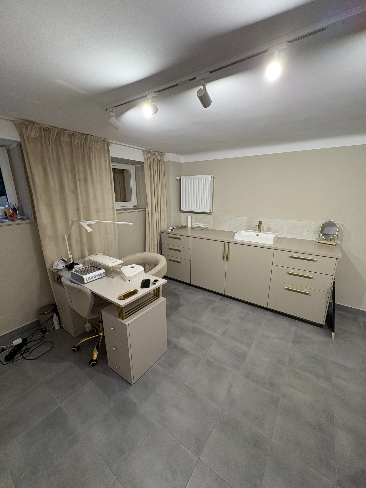 Salon kosmetyczny z beżowymi meblami, lampą nad stanowiskiem pracy, szarymi płytkami podłogowymi i białymi światłami punktowymi na suficie.