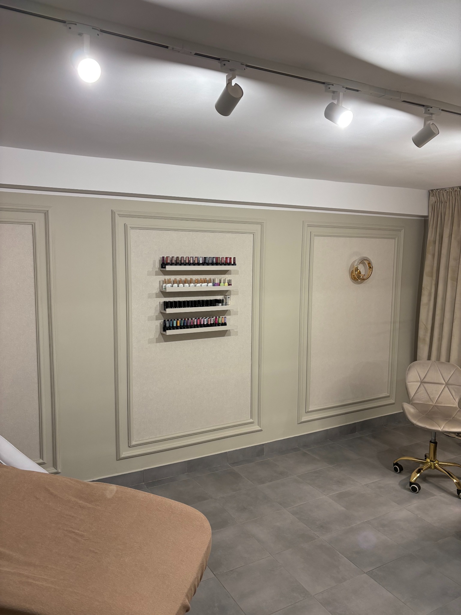 Salon kosmetyczny z beżowymi ścianami w panelach, półkami pełnymi lakierów do paznokci, regulowanym krzesłem na kółkach i lampami sufitowymi.