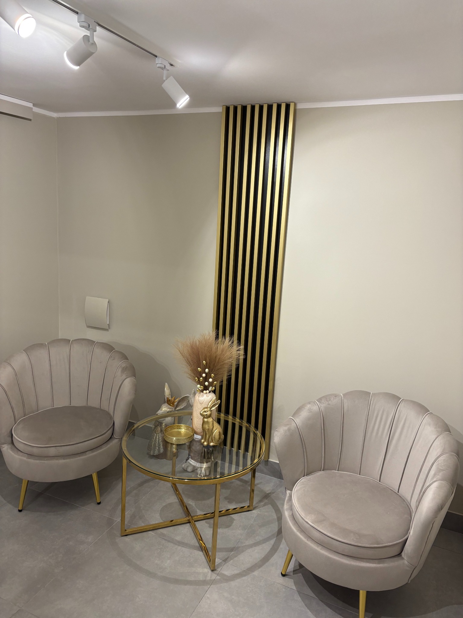 Beżowe fotele muszelki z okrągłym szklanym stolikiem na złotych nogach, dekoracje i pionowe czarno-złote panele ścienne w nowoczesnym, minimalistycznym wnętrzu z oświetleniem punktowym.
