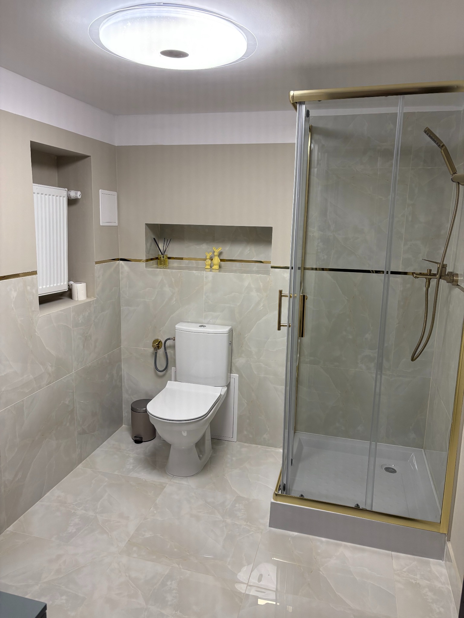 Jasna łazienka z kabiną prysznicową w złotej ramie, toaletą i dekoracyjną wnęką z oświetleniem sufitowym.