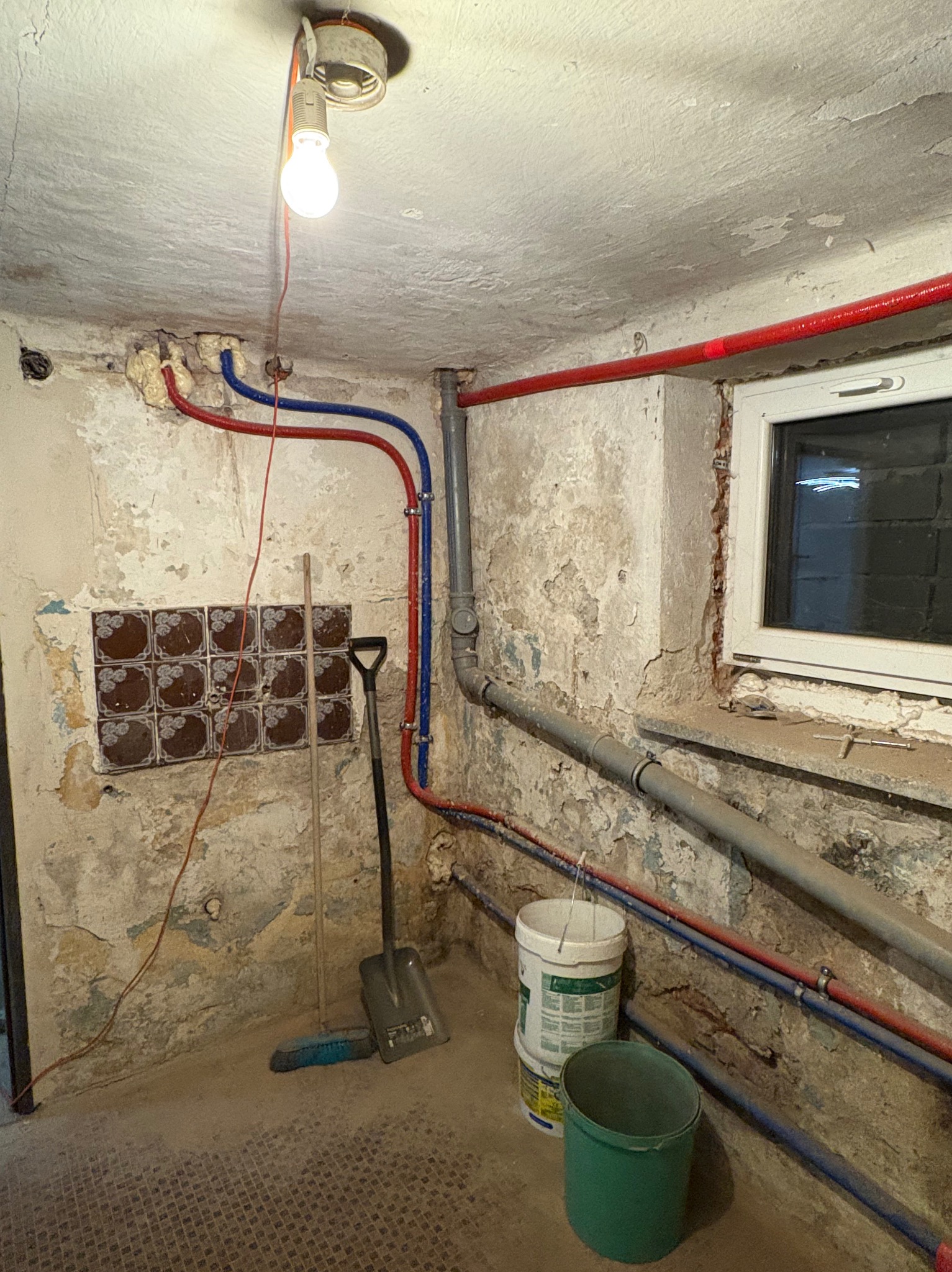 Zdjęcie zaniedbanej łazienki w trakcie renowacji: widoczne stare rury, odrapane ściany z resztkami płytek, wiadro farby i narzędzia. Żarówka oświetla wnętrze.