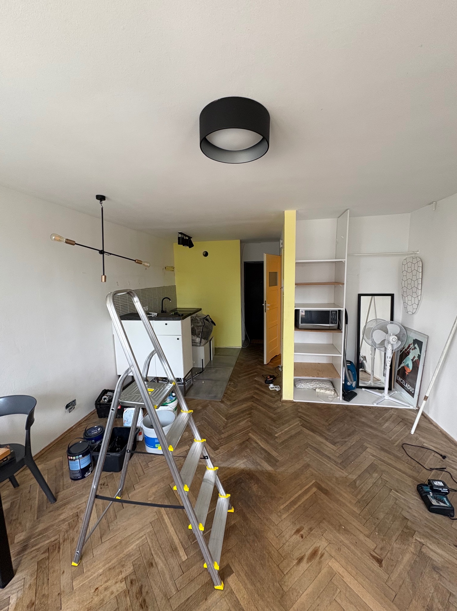 Kawalerka przed malowaniem: widok na pokój z drabiną, farbami, sprzętem AGD i meblami. Żółta ściana w tle. Parkiet w jodełkę na podłodze. Wnętrze w trakcie przygotowań do prac.
