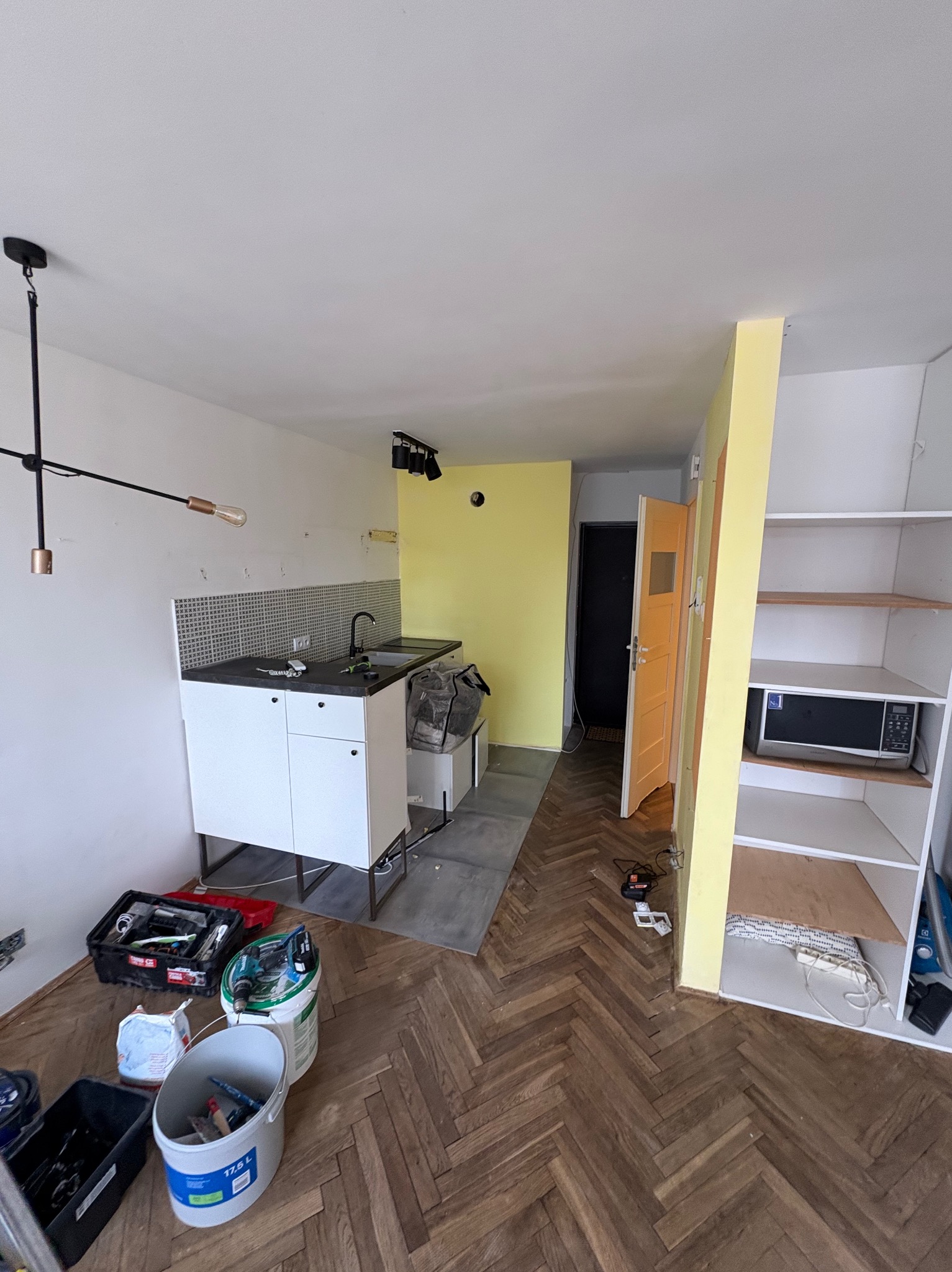 Wnętrze kawalerki przed malowaniem: widok na kuchnię z narzędziami malarskimi, żółtą ścianą i parkietem w jodełkę. Białe szafki kuchenne i regał z mikrofala.