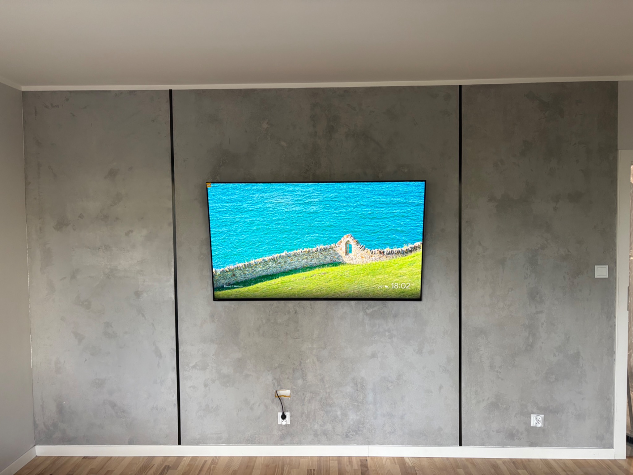 Ściana w odcieniach szarości z dekoracyjną fakturą, podzielona czarnymi listwami. Na ścianie zawieszony telewizor. Widoczne gniazdka elektryczne i drewniana podłoga.