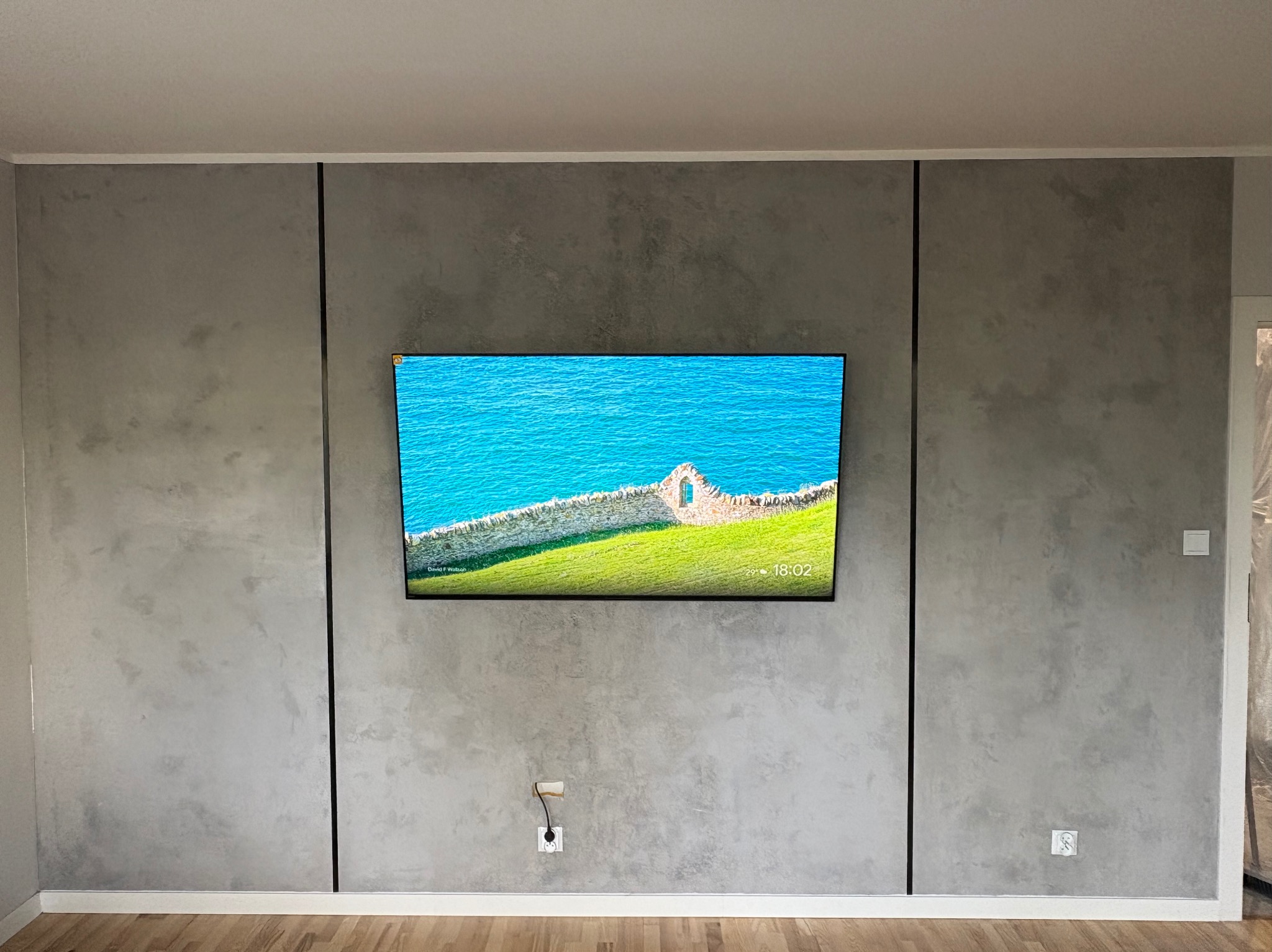 Szara ściana z efektem betonu, podzielona czarnymi listwami, z wiszącym telewizorem. Widoczna drewniana podłoga i gniazdko elektryczne.