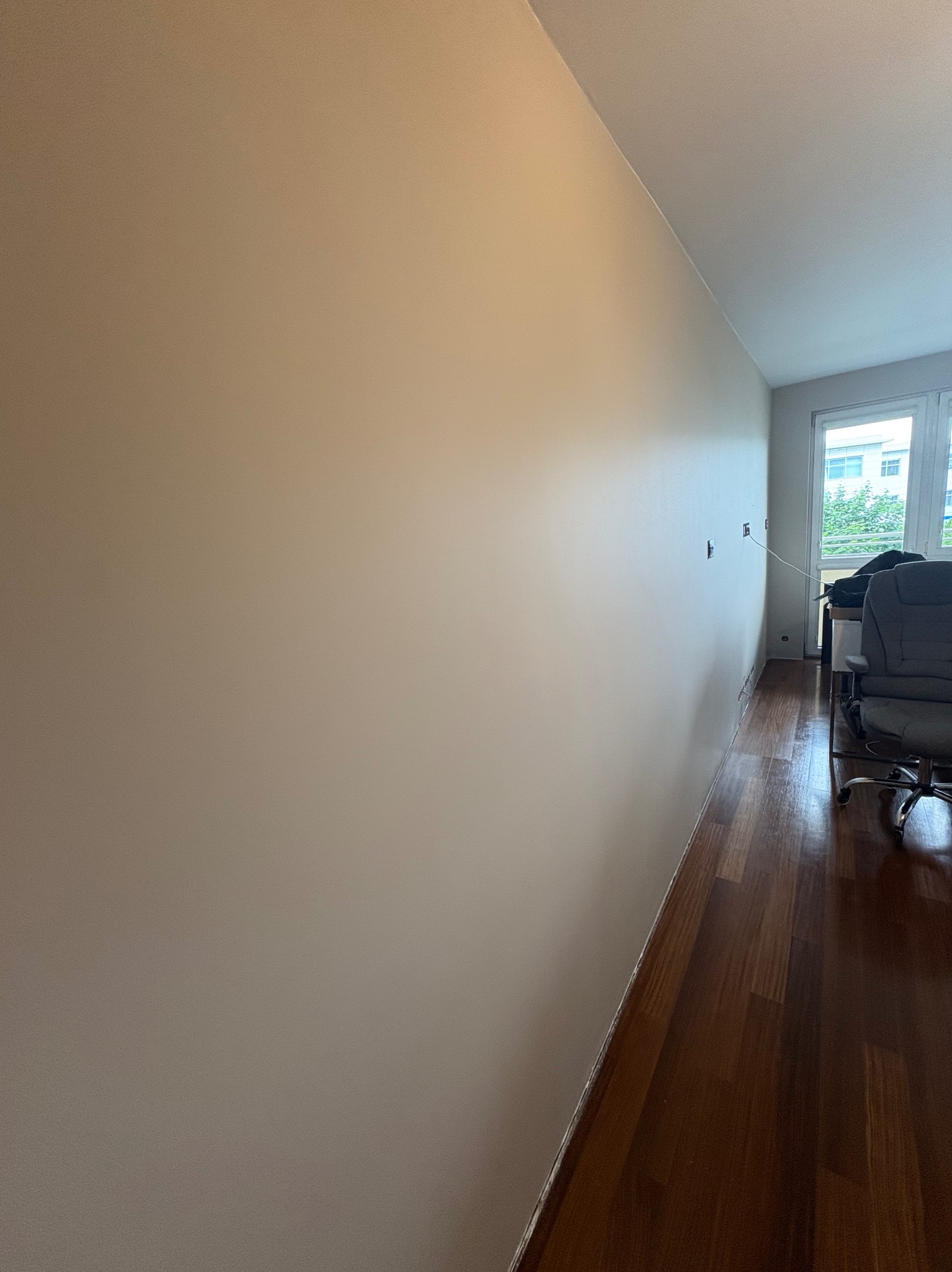 Ściana w odcieniach beżu po malowaniu, widoczne gniazdka elektryczne, parkiet i fragment biura z krzesłem w tle, neutralne oświetlenie.