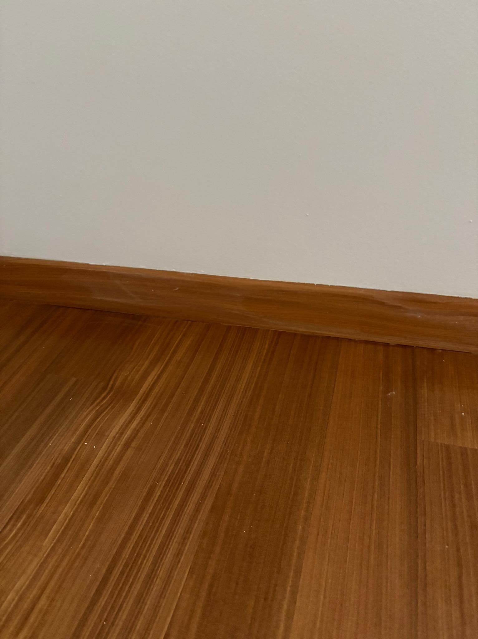 Brązowa podłoga z paneli laminowanych i pomalowana na biało ściana z brązową listwą przypodłogową. Ujęcie pod kątem, widoczne drobne zabrudzenia na podłodze.