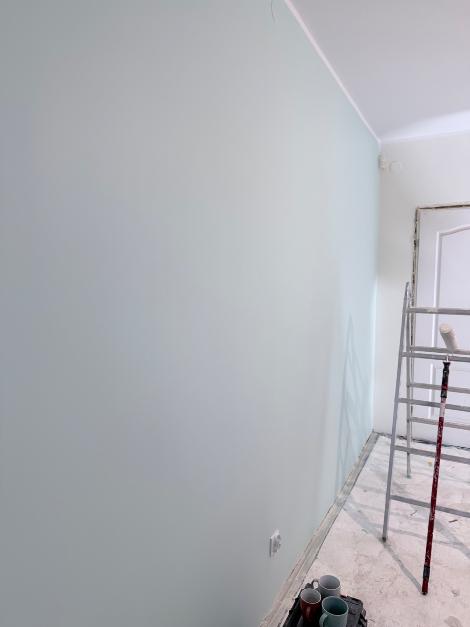 Świeżo pomalowana ściana na pastelowy odcień z widoczną drabiną i wałkiem malarskim. W rogu podłogi widoczne kubki i skrzynka narzędziowa.