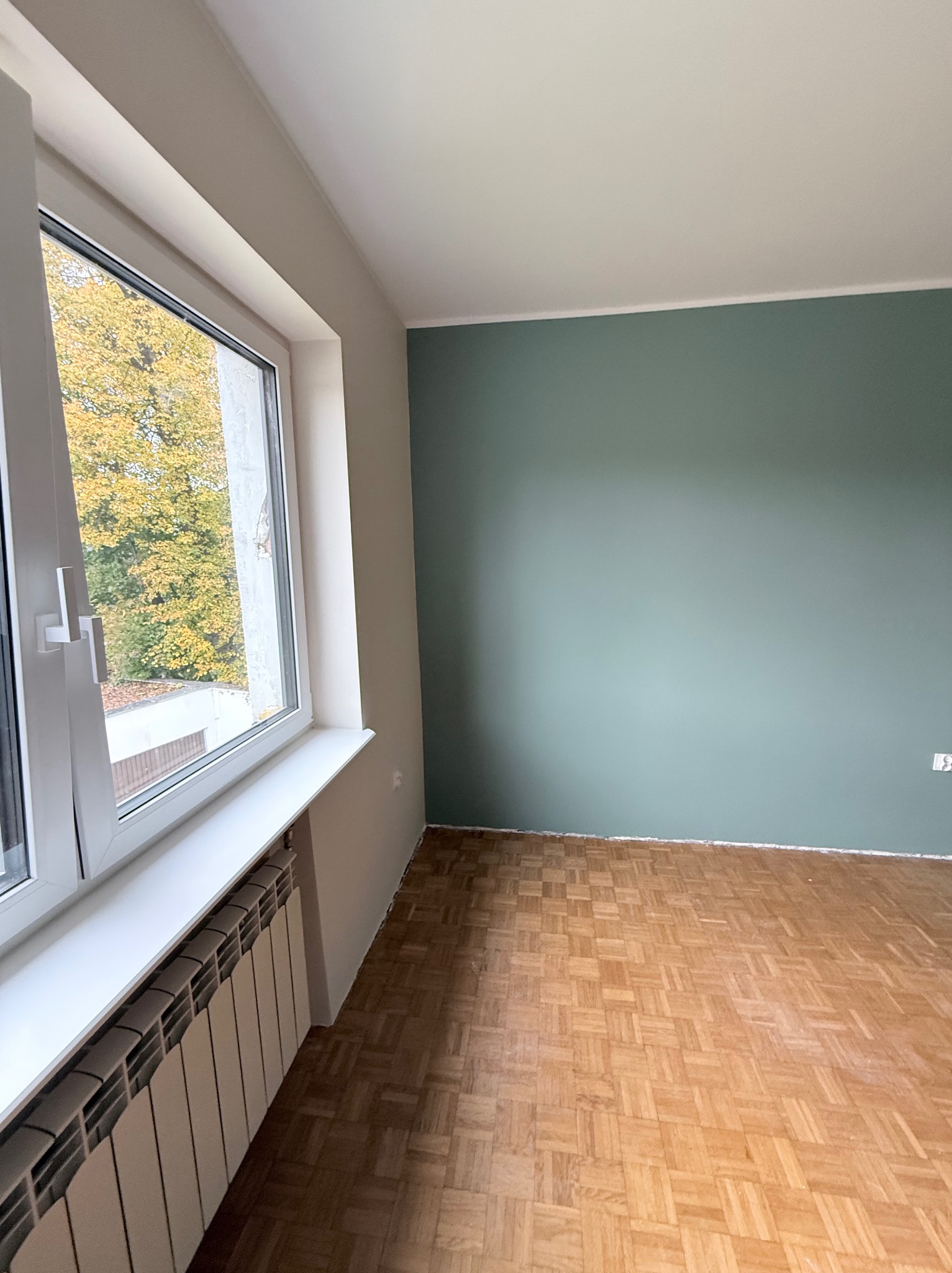 Pomieszczenie po malowaniu ścian na kolor morski, z widocznym parkietem, grzejnikiem i oknem z widokiem na drzewa z żółtymi liśćmi. Ściany pomalowane do połowy wysokości.