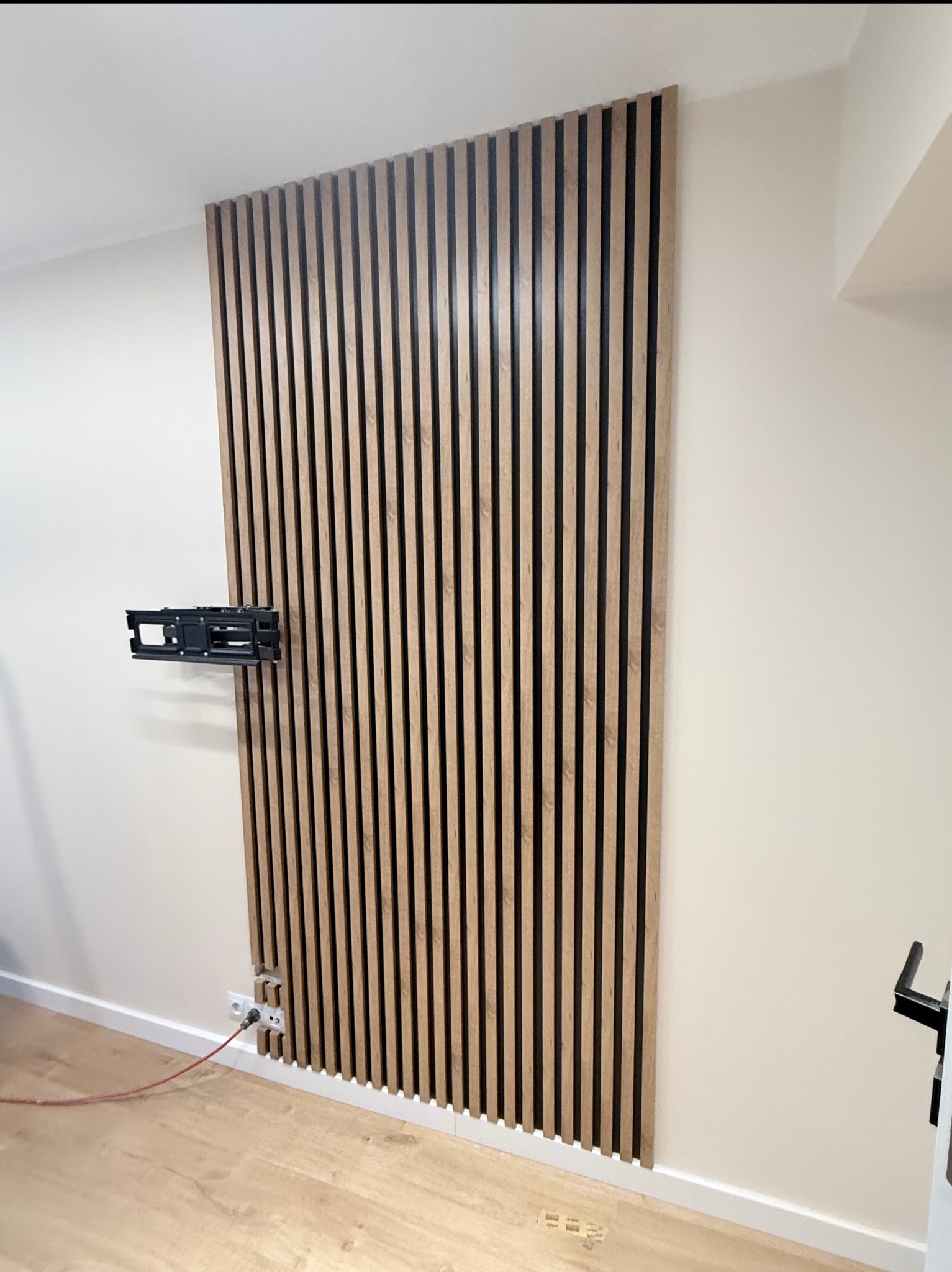 Panel lamelowy z drewna na ścianie, z zamontowanym uchwytem na telewizor i gniazdkiem elektrycznym wkomponowanym w konstrukcję. Jasne ściany i podłoga.