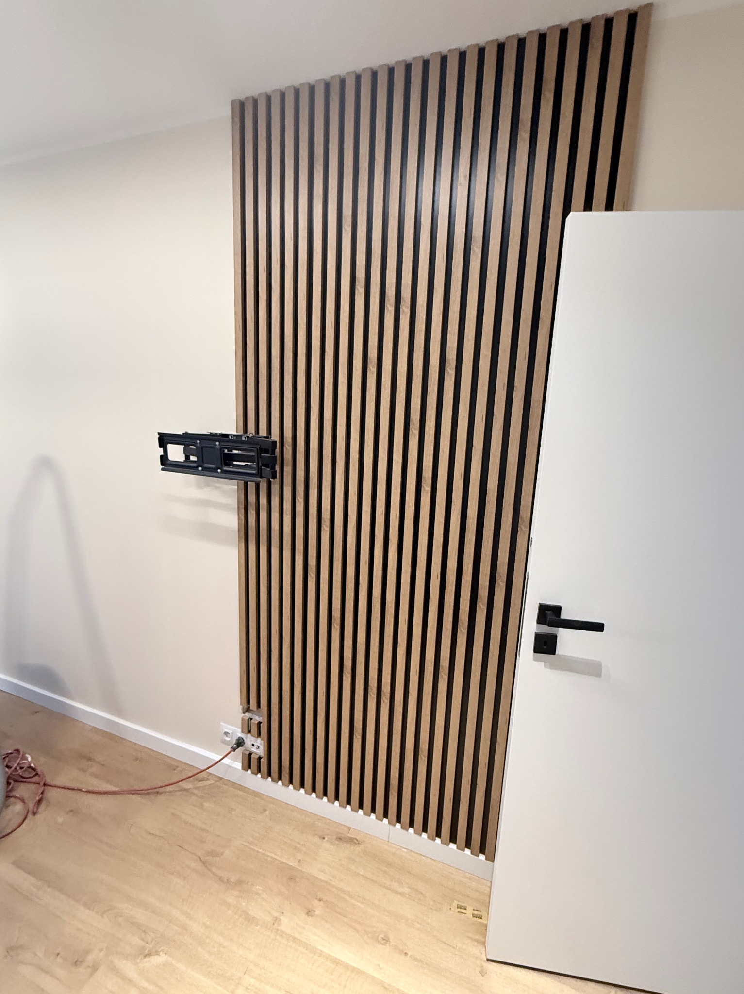 Panel dekoracyjny z drewnianych listew na ścianie, obok białe drzwi z czarną klamką. Widoczny uchwyt na telewizor i gniazdko elektryczne. Podłoga drewniana.