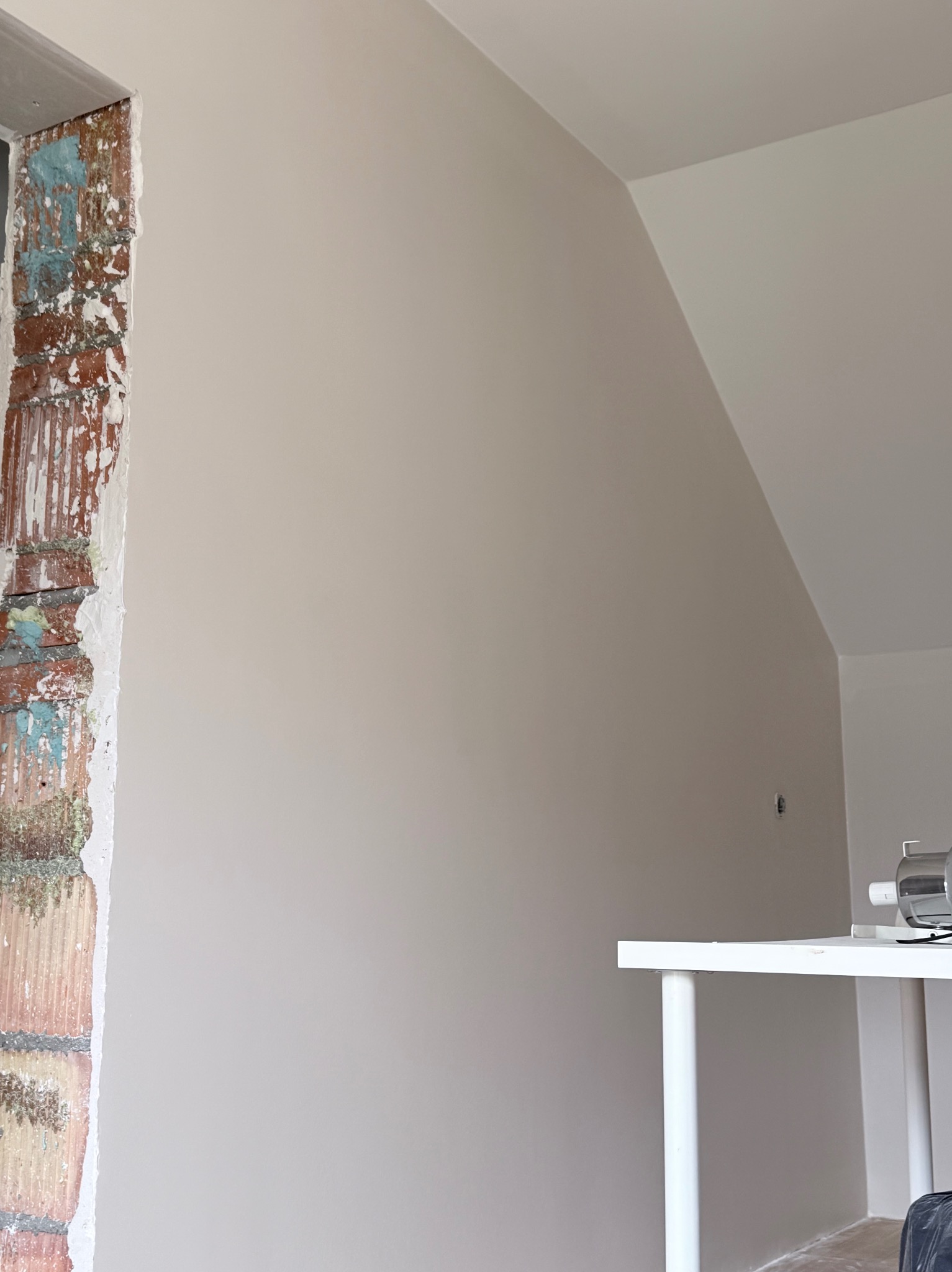 Ściana po malowaniu na neutralny odcień, z fragmentem odsłoniętej cegły przy oknie. Widoczny fragment białego stolika i skos dachu.