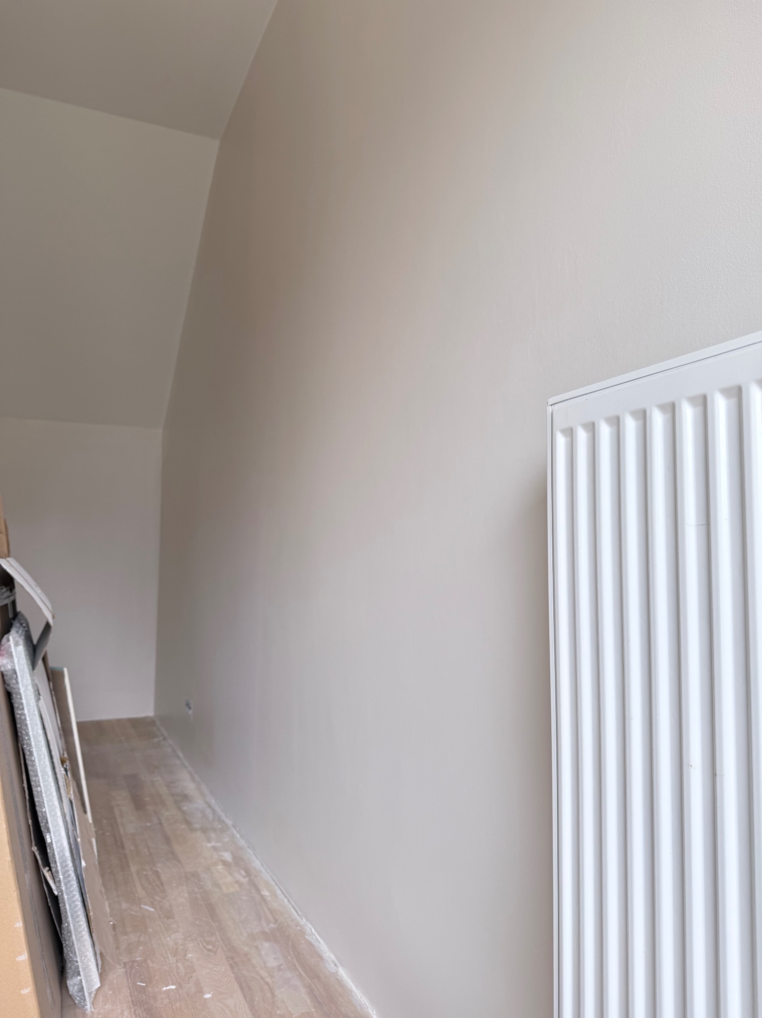 Jasne wnętrze poddasza po malowaniu ścian na beżowo, z widocznym białym grzejnikiem i fragmentem podłogi. W rogu leżą zabezpieczone folią ramy obrazów.
