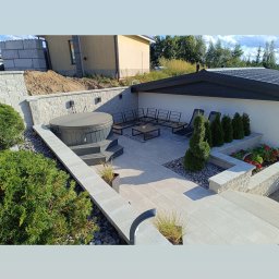 Zbigniew Choszcz - Nowoczesny taras z szarej kostki, jacuzzi, meblami ogrodowymi i roślinami w donicach. Kamienne murki oddzielają strefy relaksu. Widok z góry.