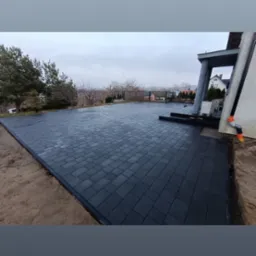 Rozległy taras z ciemnej kostki brukowej, obramowany piaskiem, przylegający do nowoczesnego domu z widokiem na zalesione wzgórze w tle.