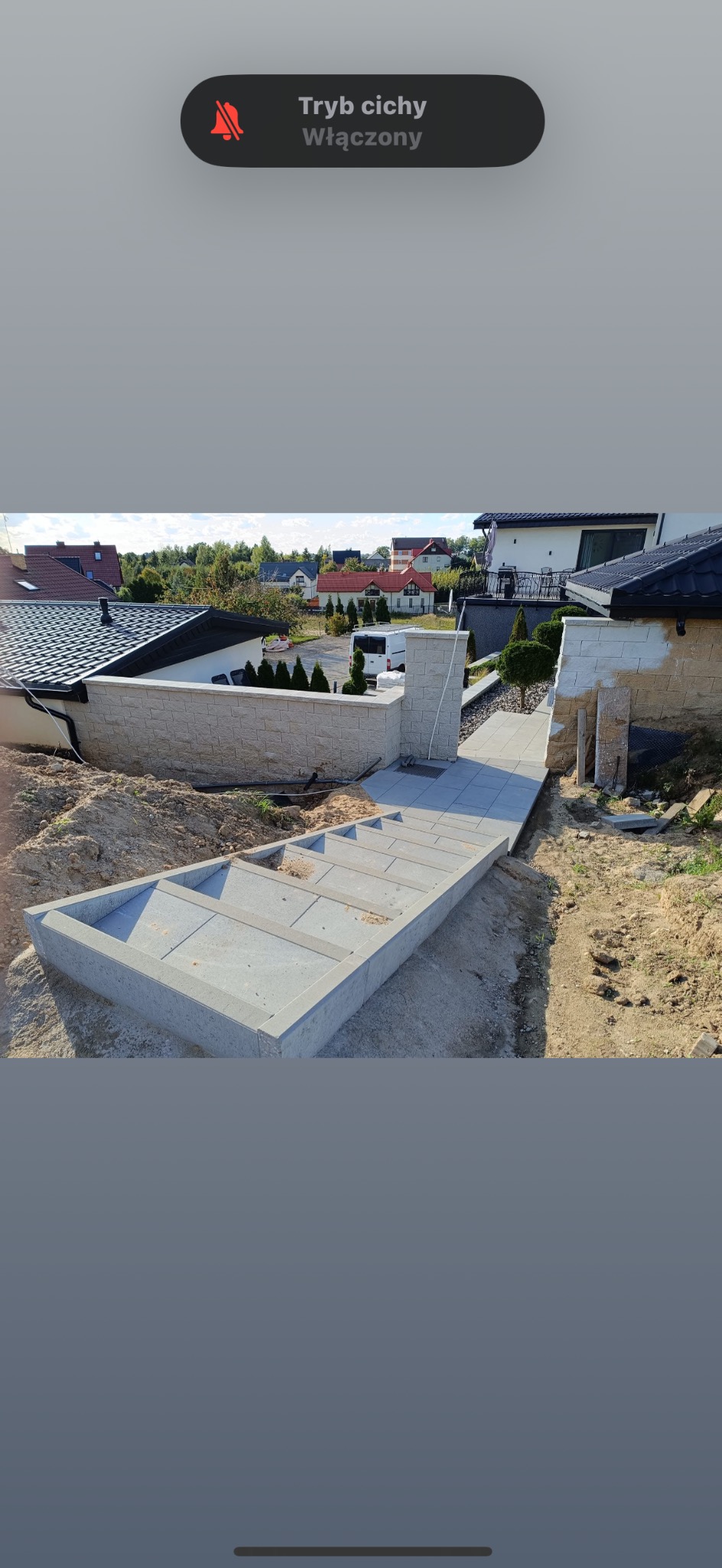 Schody z szarej kostki prowadzące do domu z murem z jasnego kamienia. Częściowo ułożona kostka, widoczne prace ziemne. Tło stanowią domy i zieleń.