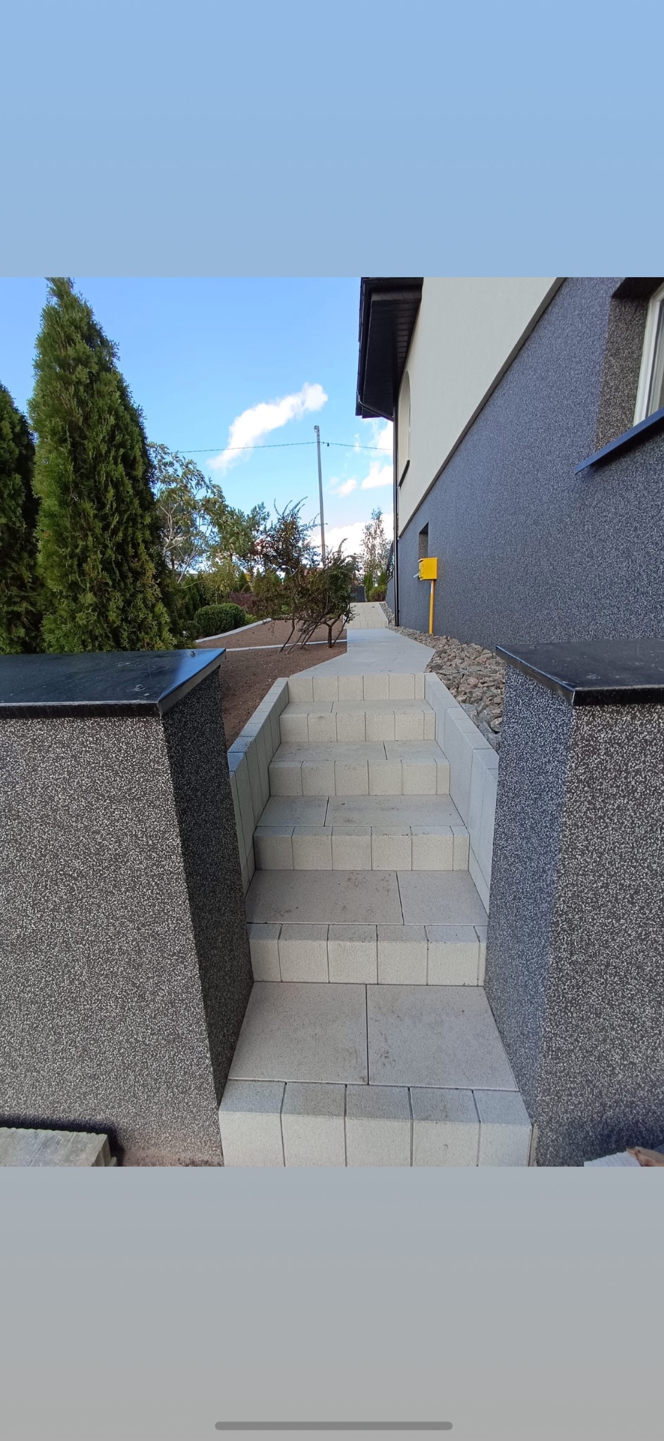 Schody z jasnej kostki brukowej prowadzące do domu z szarą elewacją, otoczone zielenią i elementami dekoracyjnymi z ciemnej kostki.