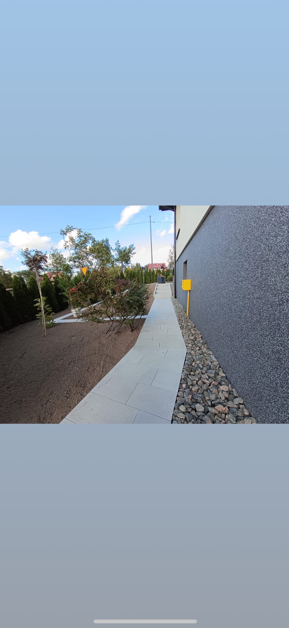 Jasnoszara ścieżka z kostki brukowej prowadzi wzdłuż ściany budynku z szarą elewacją. Obok ścieżki rabata z roślinami i kamieniami. Błękitne niebo w tle.