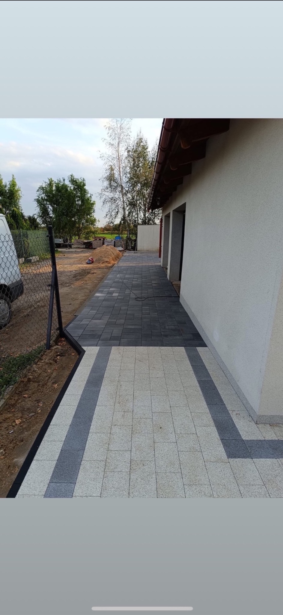 Ułożona kostka brukowa w dwóch odcieniach szarości tworząca wzór ścieżki przy budynku, obok ogrodzenie z siatki.